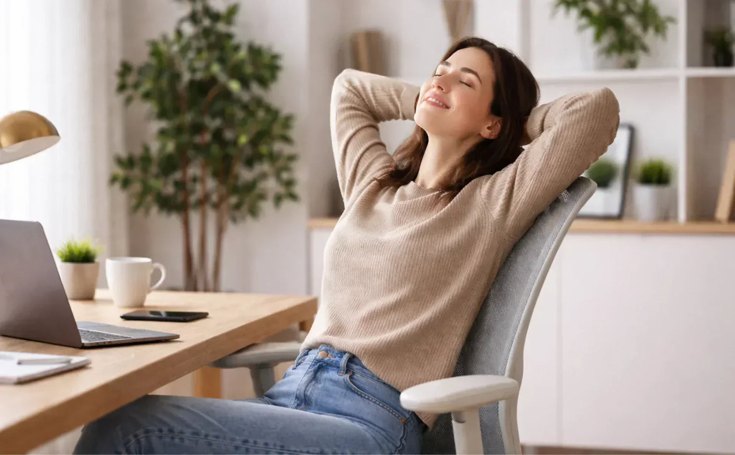 A happy woman leaning back and relaxing on a grey ergonomic mesh office chair at a wooden home office desk with a laptop.